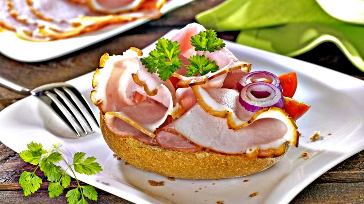 Ein belegtes Brötchen mit Schinkenscheiben, roten Zwiebeln, Tomaten und Petersilie auf einem weißen Teller mit Gabel.