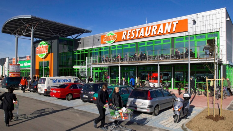 Eine GLOBUS-Filiale mit grün-orangen Logos, Parkplatz mit Autos, Einkaufswagen und Restaurant mit Balkon voller Gäste.
