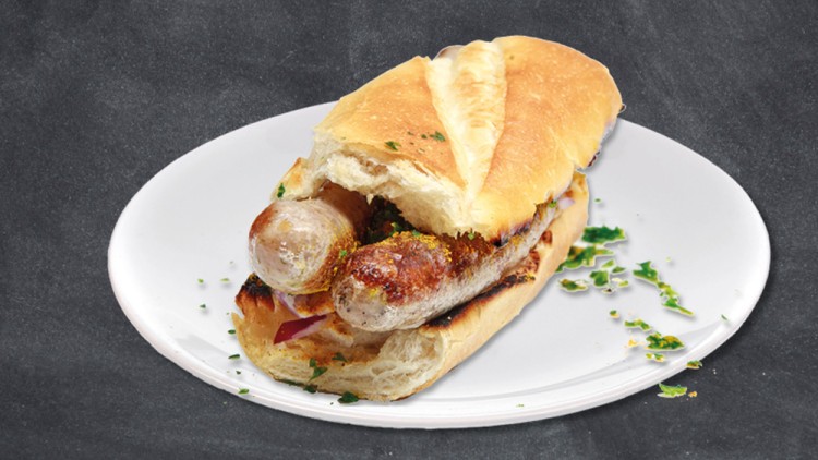 Brötchen mit zwei gebratenen, braunen Würstchen und grüner Kräuterdekoration auf weißem Teller vor grauem Hintergrund.