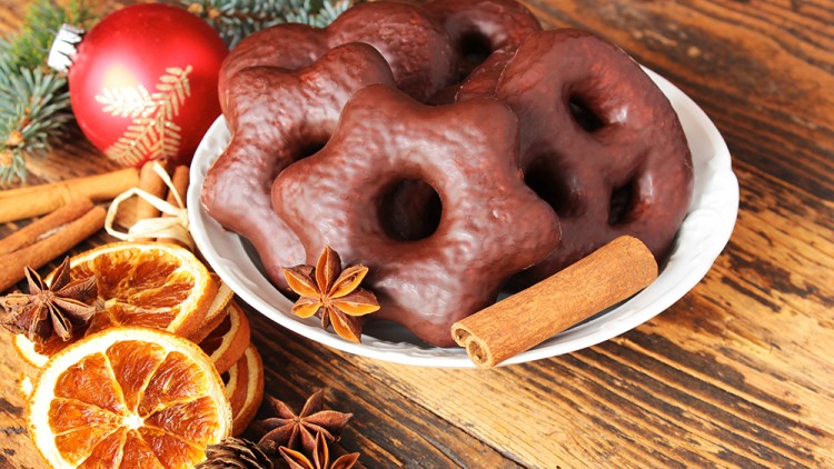 Schokoladen-Lebkuchen in Stern- und Herzform auf weißer Schale, daneben Zimtstangen, Sternanis, Orangenscheiben und rote Weihnachtskugel.