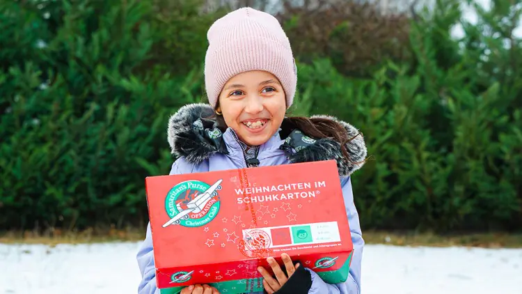 Kind in Winterkleidung hält roten Karton mit der Aufschrift „Weihnachten im Schuhkarton“ vor grünen Büschen und Schnee.
