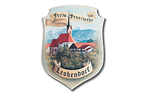 Ein Wappen mit einer Kirche vor Bergen, umrahmt von Bäumen und zwei Bändern mit der Aufschrift Freiw Feuerwehr Leobendorf.