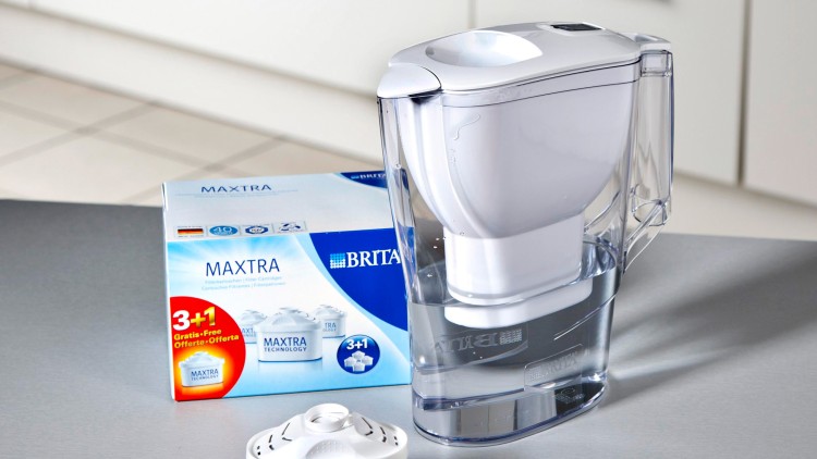 Ein durchsichtiger Wasserfilterkrug mit weißem Deckel steht neben einer Verpackung und einem Ersatzfilter der Marke BRITA MAXTRA.