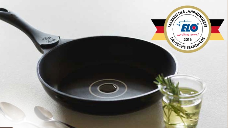 Schwarze Pfanne mit Aufschrift „ELO 1921“ neben einem Glas mit Öl und Rosmarin, drei Löffeln und zwei Auszeichnungen abgebildet.