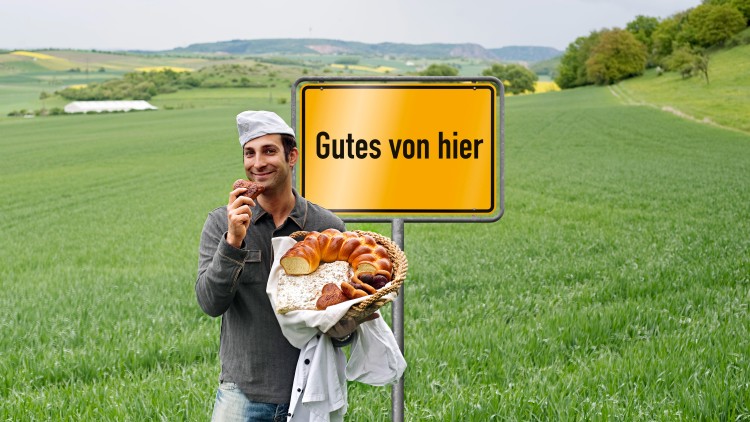 Person hält einen Korb mit frischen Backwaren vor einem gelben Schild mit der Aufschrift Gutes von hier, grüne Wiese im Hintergrund.