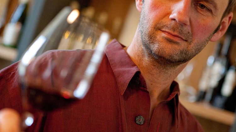 Ein Mensch in einem weinroten Hemd hält ein Glas mit Rotwein vor einem Regal mit Weinflaschen.