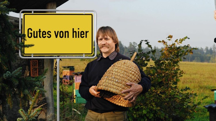 Eine Person hält einen traditionellen Korb, im Hintergrund ein Schild mit der Aufschrift Gutes von hier und eine grüne Landschaft.
