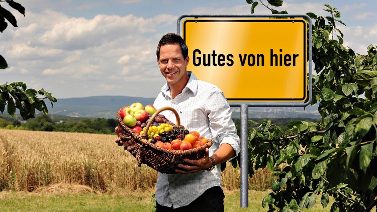 Ein Korb mit Obst wie Äpfeln, Trauben und Pfirsichen vor einem gelben Schild mit der Aufschrift Gutes von hier in ländlicher Umgebung.