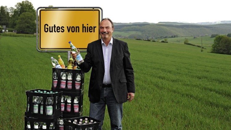 Mann in Anzug hält Mineralwasserflasche vor grünem Feld, gelbes Schild mit Aufschrift Gutes von hier, Getränkekisten davor.