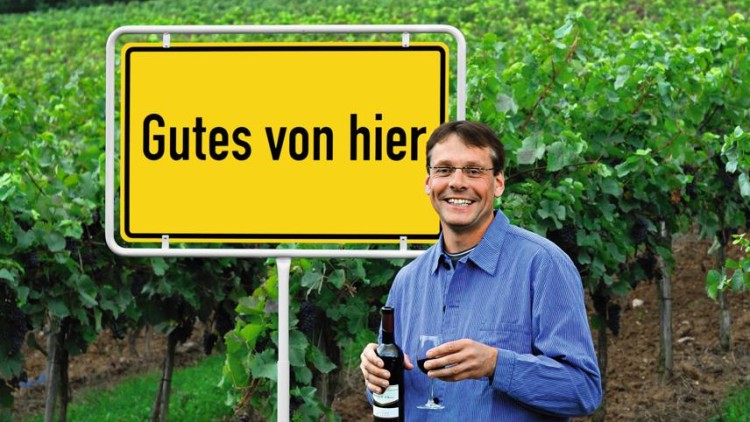 Ein gelbes Schild mit der Aufschrift „Gutes von hier“ steht vor einem Weinberg, daneben eine Person mit Weinflasche und Glas.