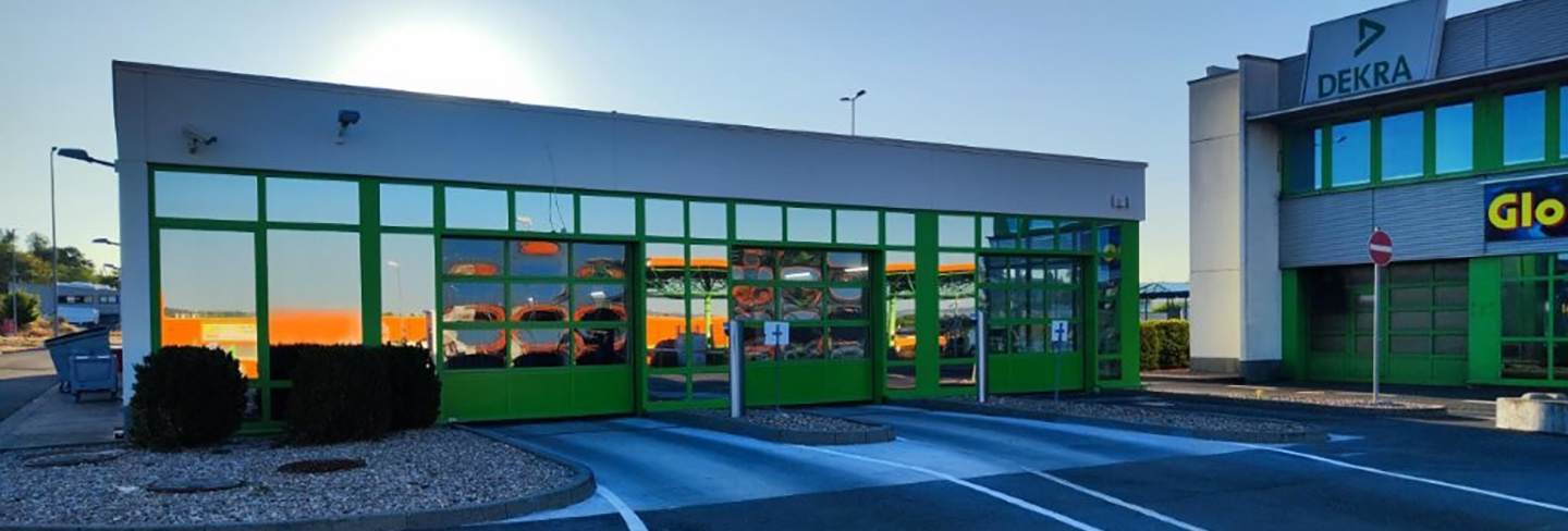 Gebäude mit großer Glasfront, grünen Rahmen und angrenzendem DEKRA-Gebäude, Parkplatz mit markierten Stellflächen davor.
