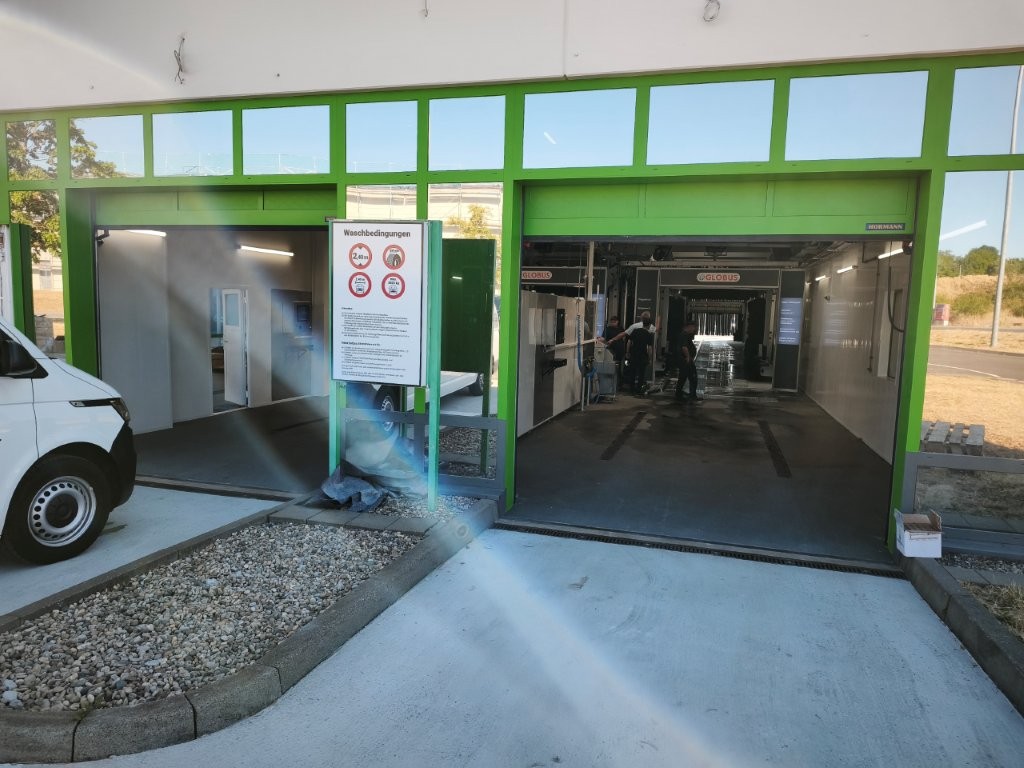 Grüne Autowaschanlage mit zwei Einfahrten, Waschanweisungen auf einem Schild und Mitarbeiter bei der Fahrzeugreinigung.