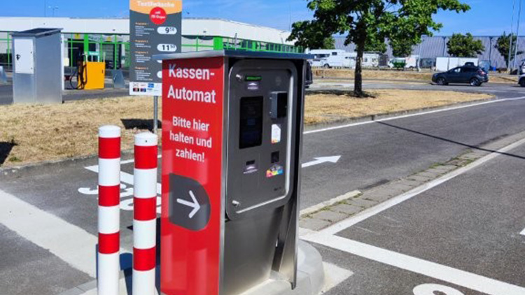 Roter Kassenautomat mit der Aufschrift „Bitte hier halten und zahlen“, daneben rot-weiß gestreifte Poller, Parkplatz im Hintergrund.