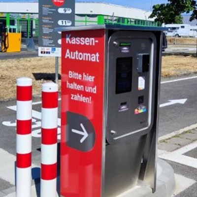 Roter Kassenautomat mit der Aufschrift „Bitte hier halten und zahlen“, daneben rot-weiß gestreifte Poller, Parkplatz im Hintergrund.