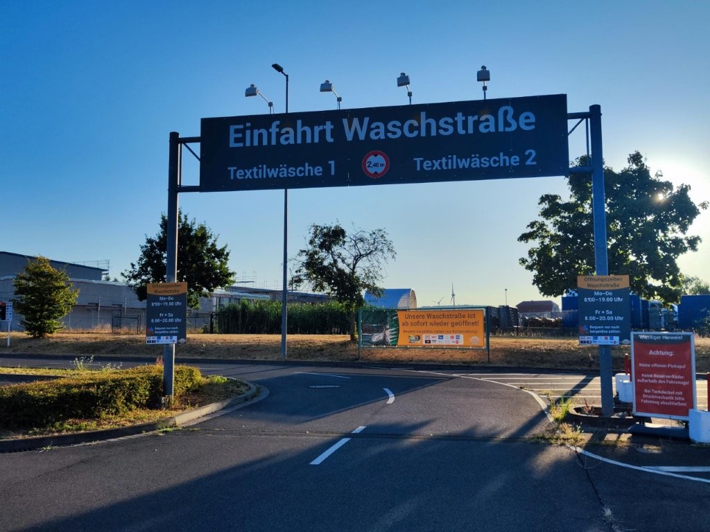 Schwarzes Schild mit weißer Schrift: „Einfahrt Waschstraße“, darunter „Textilwäsche 1“ und „Textilwäsche 2“ bei einer Zufahrt.