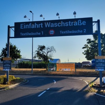 Schwarzes Schild mit weißer Schrift: „Einfahrt Waschstraße“, darunter „Textilwäsche 1“ und „Textilwäsche 2“ bei einer Zufahrt.