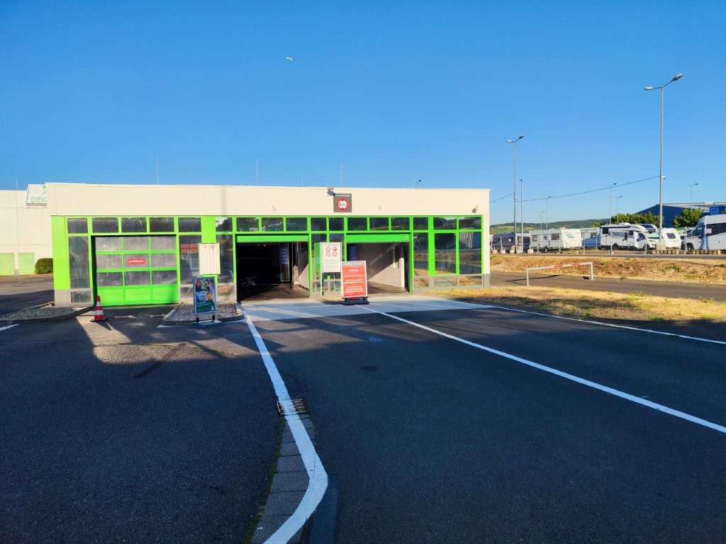 Grünes Gebäude mit Eingang und Durchfahrtsstraße, Hinweisschilder und Parkplatz mit geparkten Wohnmobilen im Hintergrund.