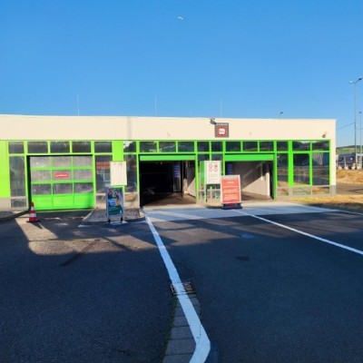 Grünes Gebäude mit Eingang und Durchfahrtsstraße, Hinweisschilder und Parkplatz mit geparkten Wohnmobilen im Hintergrund.