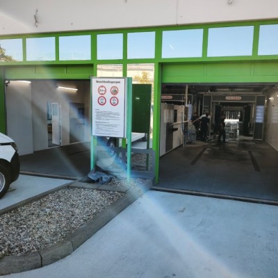 Grüne Autowaschanlage mit zwei Einfahrten, Waschanweisungen auf einem Schild und Mitarbeiter bei der Fahrzeugreinigung.