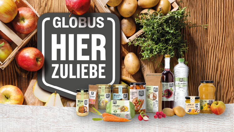 GLOBUS HIER ZULIEBE auf Schild, darunter verschiedene Lebensmittel wie Gemüse, Säfte, Kartoffeln, Äpfel und Kräuter auf hellem Untergrund.