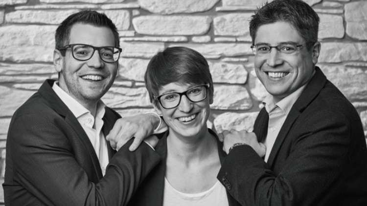Drei Personen in Anzugkleidung stehen vor einer gemusterten Steinwand und legen ihre Hände auf die Schultern der mittleren Person.