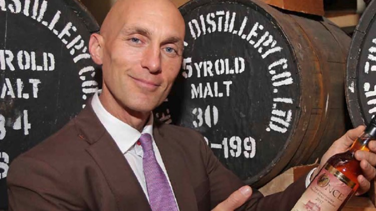 Mann in braunem Anzug hält eine Whiskyflasche, im Hintergrund stehen Holzfässer mit weißer Schrift.