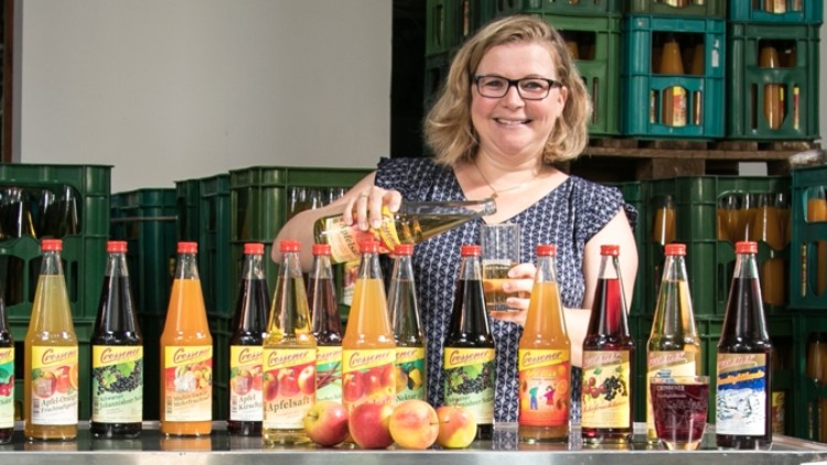 Mehrere Flaschen mit unterschiedlichem Saft stehen auf einem Tisch, davor liegen Äpfel, im Hintergrund grüne Kisten.