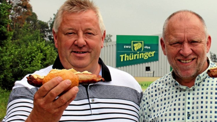 Eine Person hält eine Thüringer Rostbratwurst im Brötchen, im Hintergrund ein Schild mit der Aufschrift Thüringer EWU 1894.