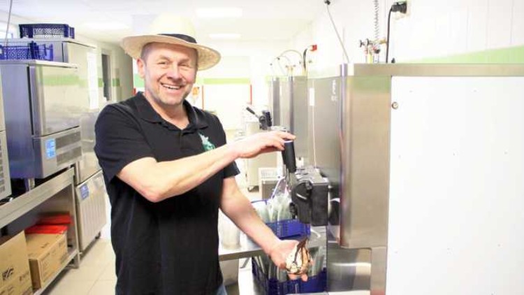 Person in schwarzem Poloshirt und Strohhut zapft Softeis aus einem silbernen Automaten in eine Waffel.