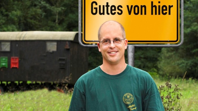 Ein gelbes Schild mit schwarzem Schriftzug „Gutes von hier“ vor einem Waggon und einer Wiese, Person im grünen Shirt im Vordergrund.