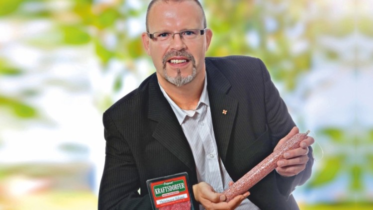 Person in dunklem Sakko hält eine längliche Salami mit rotem Etikett, daneben eine Verpackung mit der Aufschrift „Kraftsorfer“.