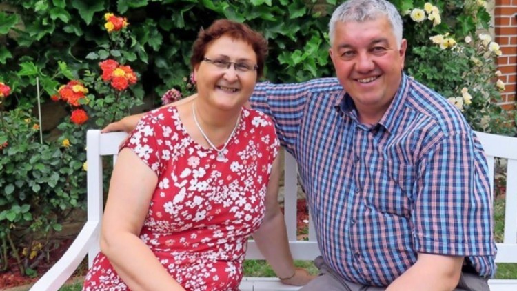 Eine Person in rotem Kleid mit Blumenmuster und eine Person in kariertem Hemd sitzen auf einer weißen Bank im Garten.