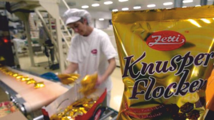 Goldene „Knusper Flocken“-Verpackung von Zetti im Vordergrund, Produktionshalle mit Förderband und Mensch im Hintergrund.