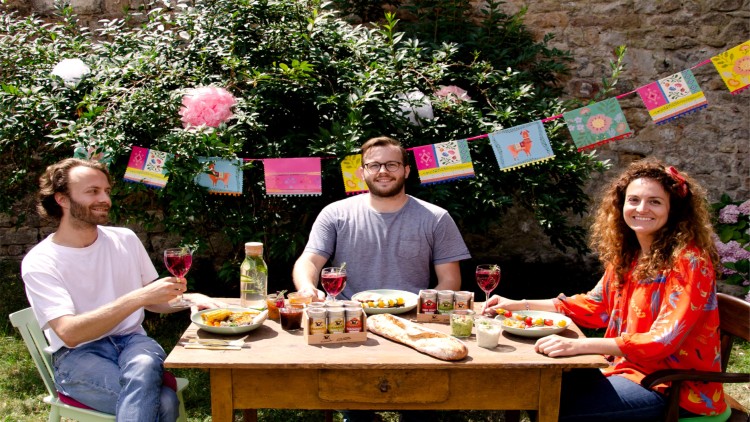 Drei Personen sitzen an einem Holztisch mit Lebensmitteln, Getränken, Brot und Dips, im Hintergrund Girlanden und grüner Busch.