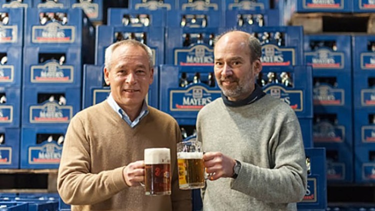 Zwei Personen halten Bierkrüge vor blauen Getränkekisten mit der Aufschrift Mayers in einer Lagerhalle.