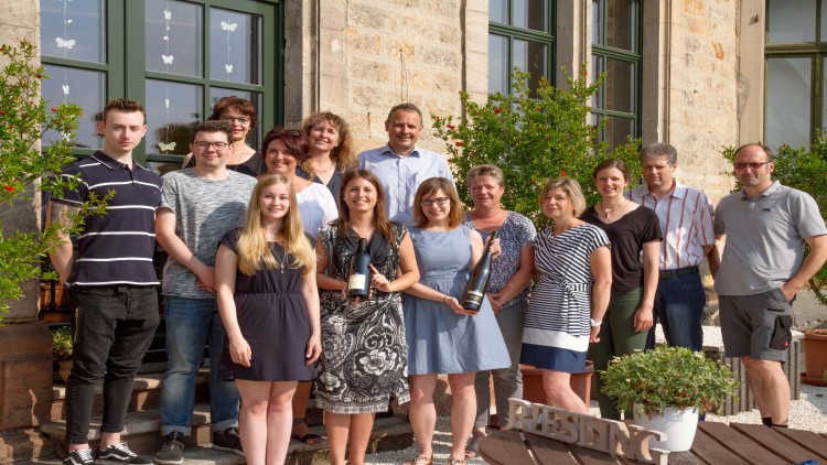 Eine Gruppe von Personen steht draußen vor einem Gebäude mit Pflanzen und einem Holzschriftzug „RIESLING“.