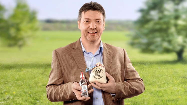 Person in braunem Sakko hält eine Glasflasche mit rotem Verschluss und ein rundes Holzprodukt mit Logo in einer Wiesenlandschaft.