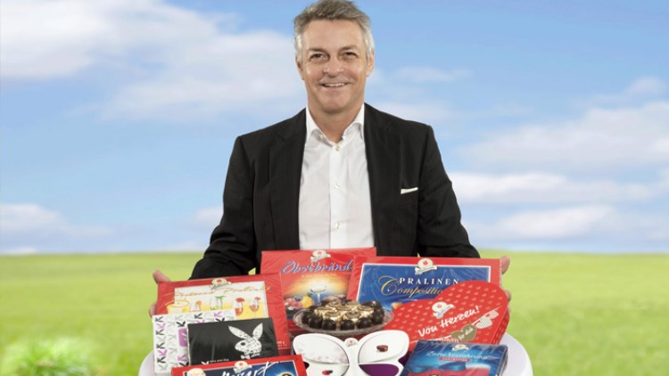 Person in Anzug hält ein weißes Tablett mit Pralinen, Schokoladenpackungen und Mozartkugeln vor einer grünen Wiesenlandschaft.