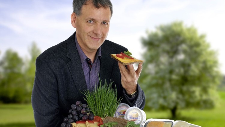 Person hält ein belegtes Brot, umgeben von frischen Lebensmitteln wie Trauben, Kräutern, Käse und Aufstrichen, im Freien.
