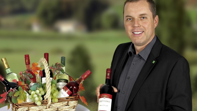 Ein Korb mit herbstlicher Dekoration und Weinflaschen steht vor einer grünen Landschaft, daneben eine Person mit einer Flasche Wein.