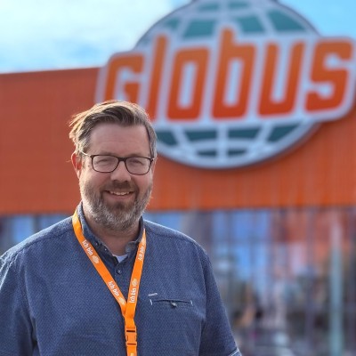 Person mit dunkelblauem Hemd, orangefarbenem Schlüsselband und Namensschild vor Eingang mit großem GLOBUS-Logo.