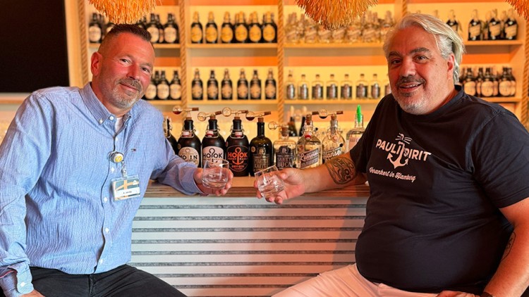Zwei Personen sitzen an einer Bar mit verschiedenen Spirituosenflaschen und Gläsern, eine trägt ein Namensschild, die andere ein schwarzes Shirt.