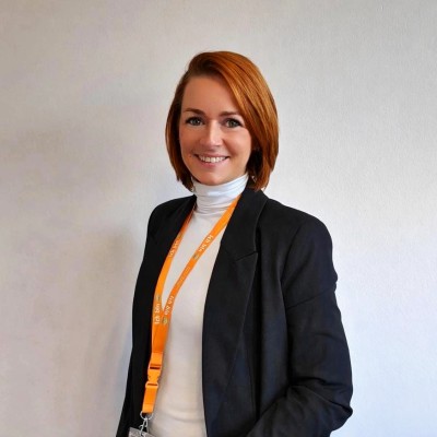 Eine Person mit rotem Haar trägt ein schwarzes Sakko, weißes Oberteil und orangefarbenes Schlüsselband vor heller Wand.