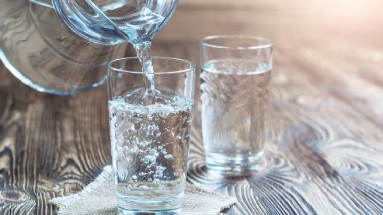 Ein Glas wird mit klarem Wasser aus einer Karaffe gefüllt, daneben steht ein weiteres Glas auf einem Holzuntergrund.