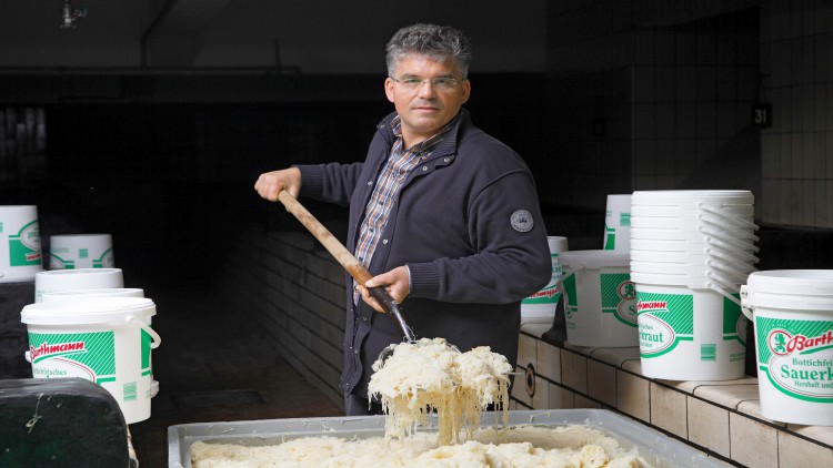 Person rührt mit Holzstab frisches Sauerkraut in einem großen Behälter um, daneben stehen weiße Eimer mit grünem Etikett.