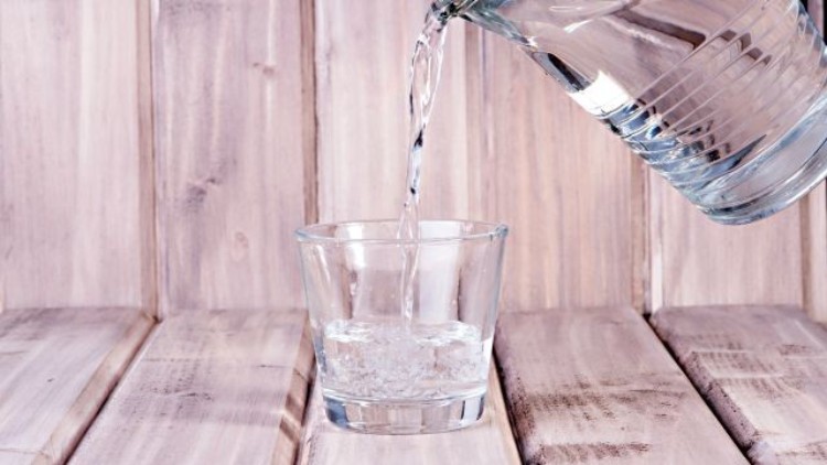 Ein durchsichtiges Glas wird mit Wasser aus einem gläsernen Krug auf einem hellen Holztisch befüllt.
