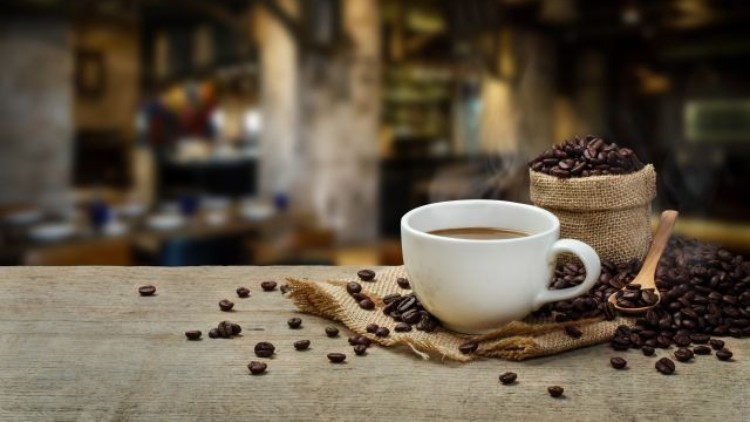 Eine weiße Tasse mit dampfendem Kaffee steht auf einem Holztisch, daneben ein Sack mit Kaffeebohnen und ein Holzlöffel.