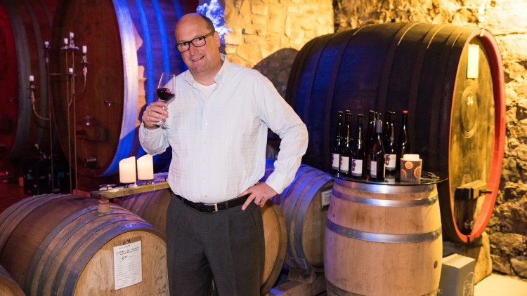 Person in weißem Hemd hält ein Glas Rotwein, umgeben von großen Holzfässern und Kerzen in einem Weinkeller.