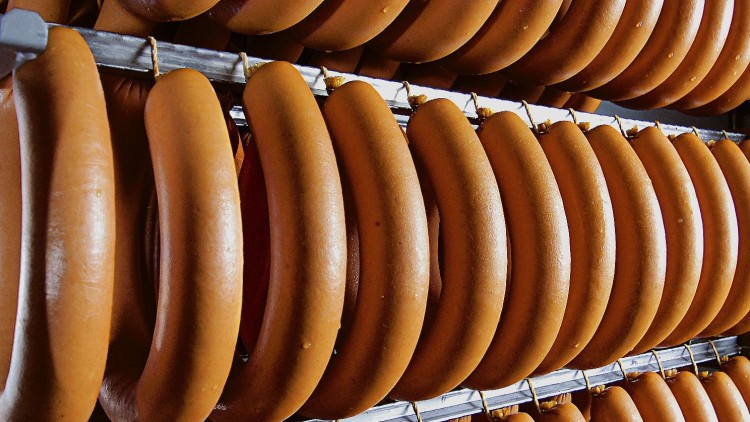 Mehrere Reihen großer brauner Fleischwürste hängen an Metallstangen in einem Lebensmittelbetrieb.