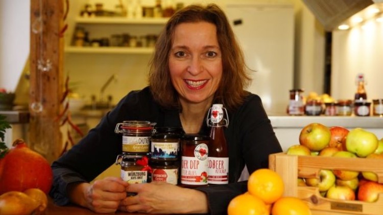 Auf einem Holztisch stehen regionale Produkte wie Marmeladen, Saucen, eine Getränkeflasche, Äpfel, Orangen und ein Kürbis.